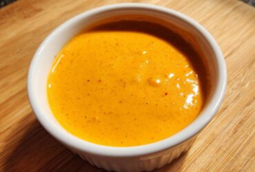 Close-up of smooth, bright orange nacho cheese sauce served in a small white ramekin on a wooden surface.