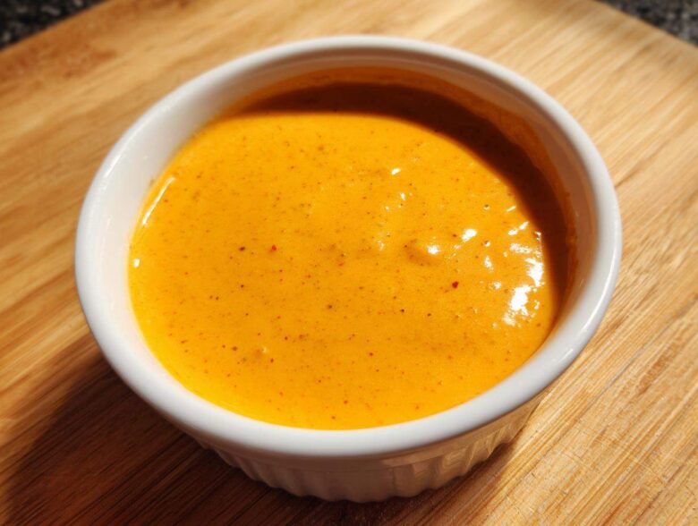 Close-up of smooth, bright orange nacho cheese sauce served in a small white ramekin on a wooden surface.