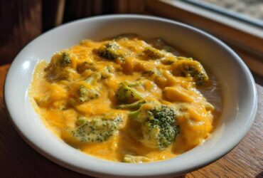 A close-up of creamy, cheesy broccoli cheddar soup served in a white bowl, highlighted by natural light.