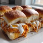 A close-up of several baked buffalo chicken sliders filled with shredded chicken, melted cheese, and drizzled with ranch dressing.