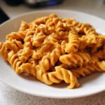 A close-up of fusilli pasta generously coated in a rich, orange butternut squash pasta sauce on a white plate.