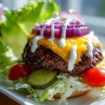 Close-up of a juicy patty topped with melted cheese, red onion, and white sauce, served over lettuce for a cheeseburger bowl.