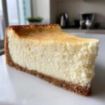 A close-up of a thick, creamy slice of baked cheesecake recipe with a golden top and graham cracker crust.