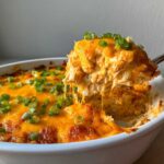 A spoonful of cheesy, shredded Buffalo chicken dip being lifted from a white baking dish, topped with green onions.