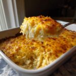 A spoonful of creamy, shredded cheesy potato casserole being lifted from a white baking dish.