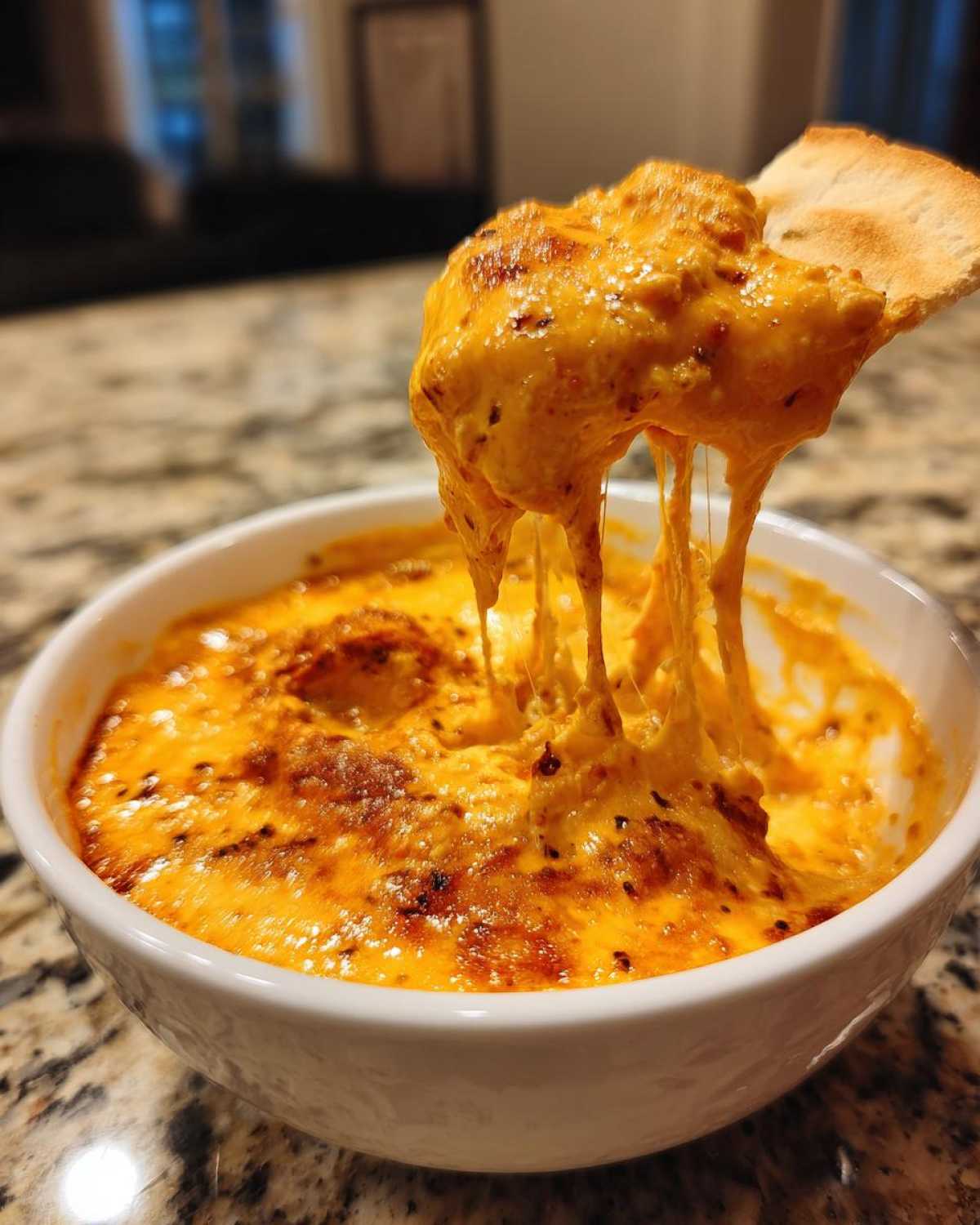 A scoop of hot, gooey chili cheese dip is lifted out of a white bowl, showing long cheese strings.