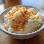 A close-up photo of creamy, white cheesy rice topped with melted orange cheddar cheese in a white bowl.