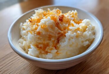 A close-up photo of creamy, white cheesy rice topped with melted orange cheddar cheese in a white bowl.