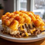 A serving of cheesy tater tot casserole showing melted cheese strings as a spoonful of ground meat filling is lifted.