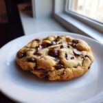 A single, thick, and perfectly baked chewy chocolate chip cookie resting on a white plate near a window.