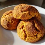 A close-up of three chewy pumpkin snickerdoodle cookies stacked on a white plate, coated in cinnamon sugar.