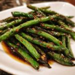A close-up of vibrant, glossy chinese green beans with garlic sauce piled on a white plate.