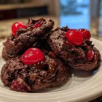 A stack of dark, rich chocolate cherry cookies topped with bright red maraschino cherries and melted chocolate chunks.