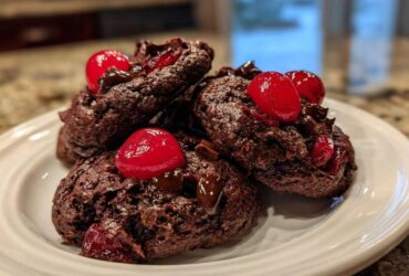 A stack of dark, rich chocolate cherry cookies topped with bright red maraschino cherries and melted chocolate chunks.