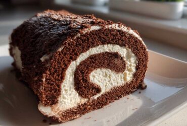 A close-up, cross-section view of a chocolate yule log cake showing the dark sponge and white cream swirl, dusted with cocoa.