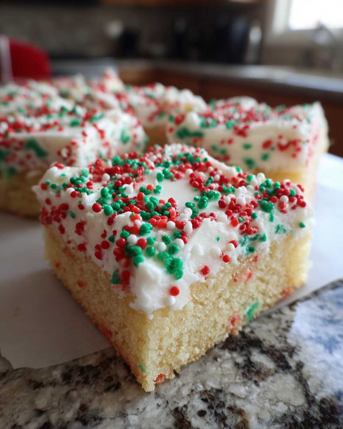 A close-up of a square slice of Christmas sugar cookie bars topped with white frosting and red and green sprinkles.