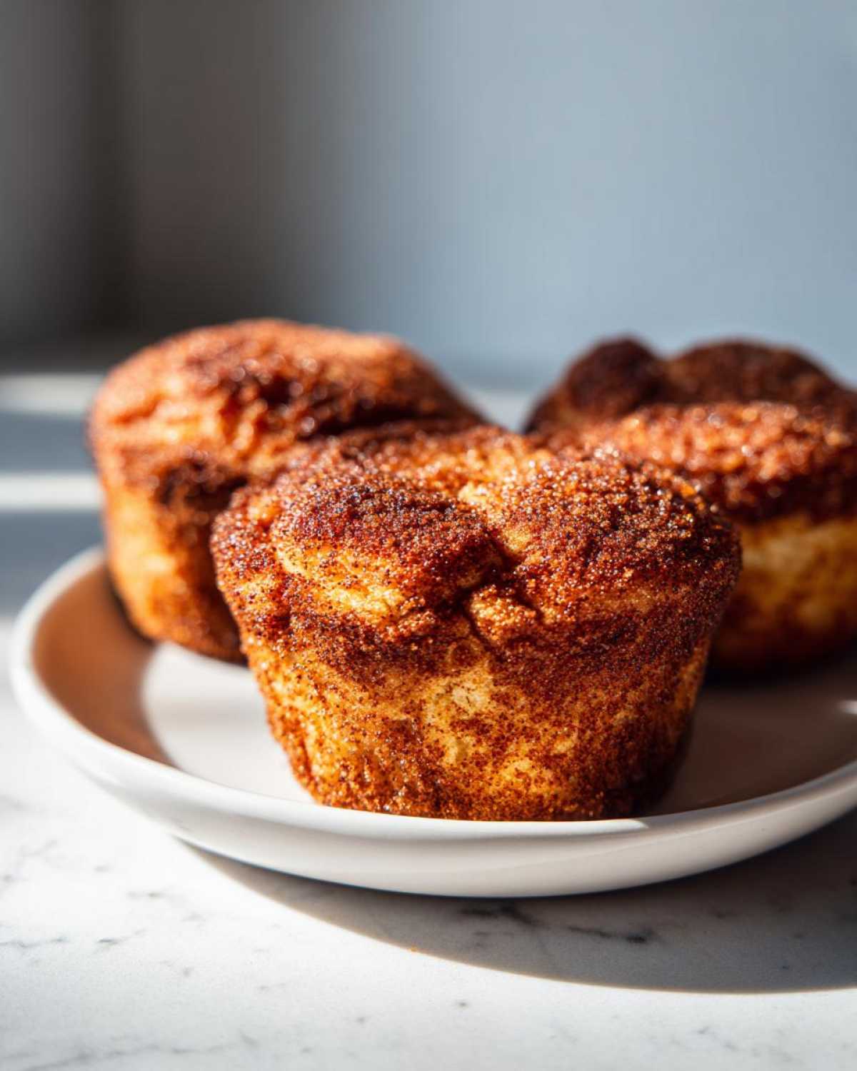 Three golden brown cinnamon sugar french toast muffins heavily coated in cinnamon sugar, sitting on a white plate.