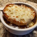 Close-up of a rich, dark french onion soup topped with a crouton and bubbly, melted Gruyère cheese.