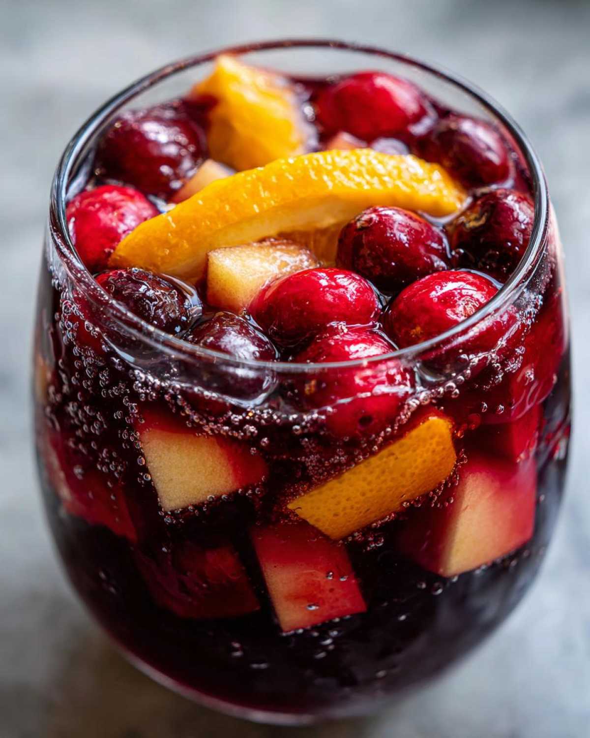 Close-up of a glass filled with bubbling red holiday sangria, loaded with cranberries, orange slices, and apple chunks.