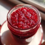 A close-up, overhead shot of thick, bright red homemade raspberry jam glistening inside a clear glass jar.