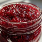 Close-up of thick, glistening homemade raspberry jam filling a small glass jar, showing texture and seeds.