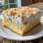 A moist slice of coconut poke cake topped with thick whipped cream and toasted coconut flakes.