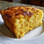 A moist, golden slice of cornbread casserole with visible corn kernels, served on a white plate.