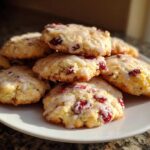 A close-up stack of freshly baked cranberry orange cookies drizzled with white glaze, sitting on a white plate.