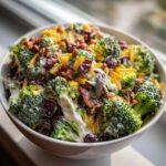 Close-up of a creamy broccoli salad topped with shredded cheddar, bacon bits, dried cranberries, and sunflower seeds.