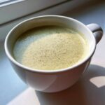 Close-up of creamy, light green celery soup with a layer of froth, served in a white mug near a window.