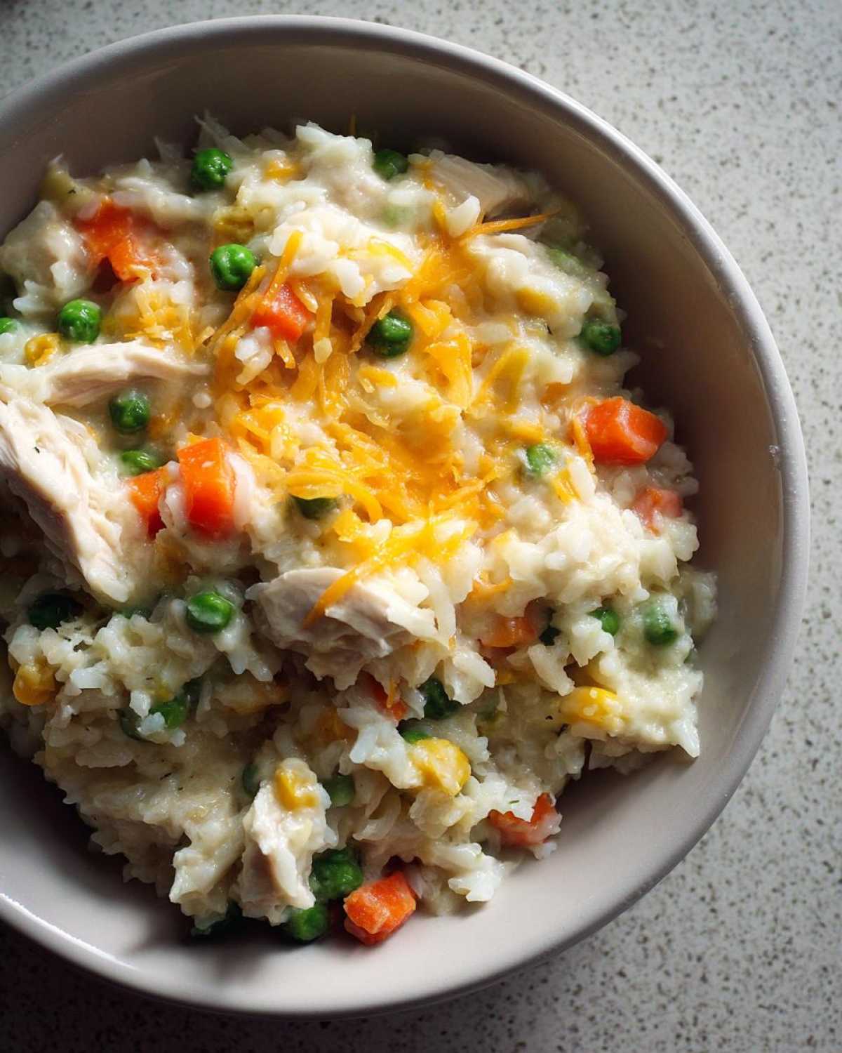 Close-up of creamy chicken and rice mixed with peas, carrots, and topped with shredded cheddar cheese.