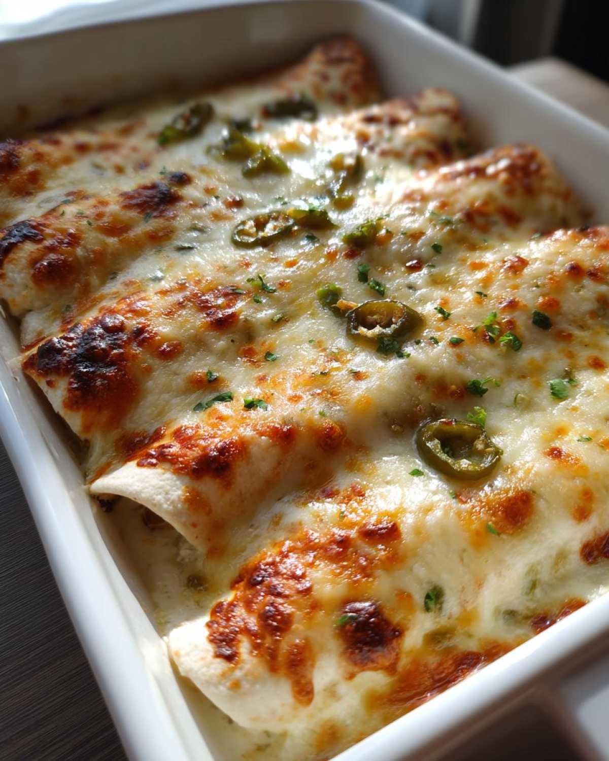 Close-up shot of creamy chicken enchiladas baked in a white dish, topped with melted, browned cheese and sliced jalapeños.