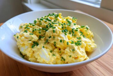 A white bowl filled with creamy, yellow egg salad recipe, generously topped with chopped green chives.