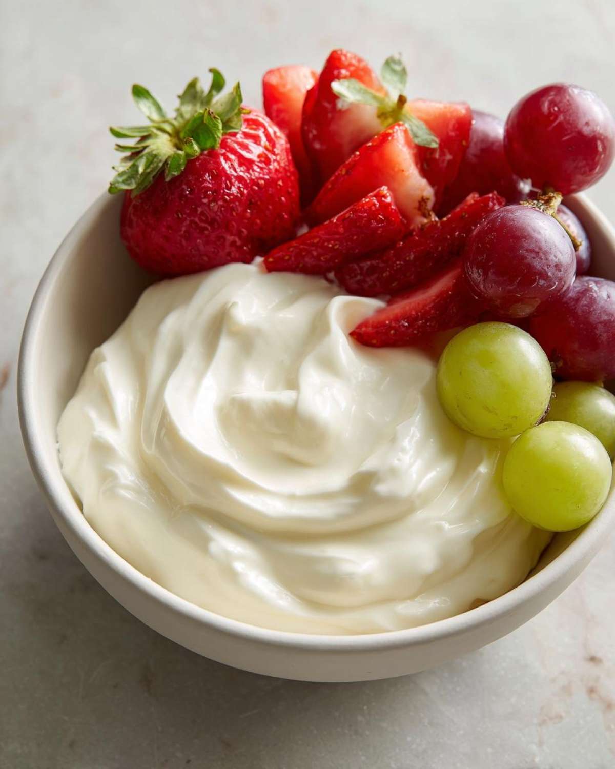 A bowl of creamy white fruit dip topped with fresh sliced strawberries and red and green grapes.