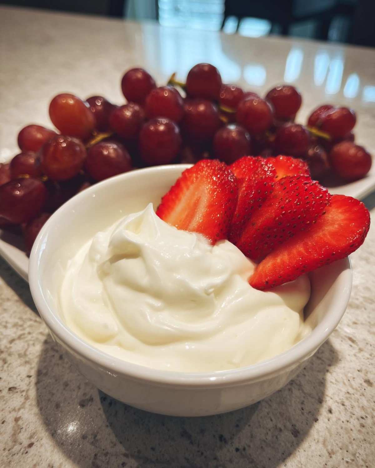 Close-up of a small white bowl filled with creamy fruit dip, topped with sliced strawberries, with red grapes in the background.