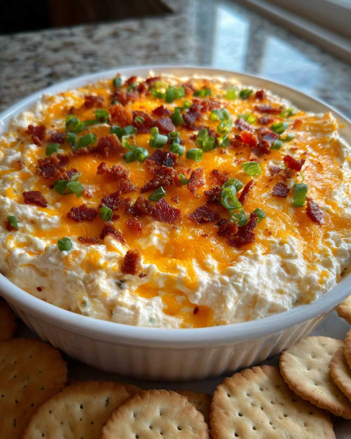 A white bowl filled with creamy Million Dollar Dip, topped with shredded cheddar cheese, bacon bits, and green onions, served with crackers.