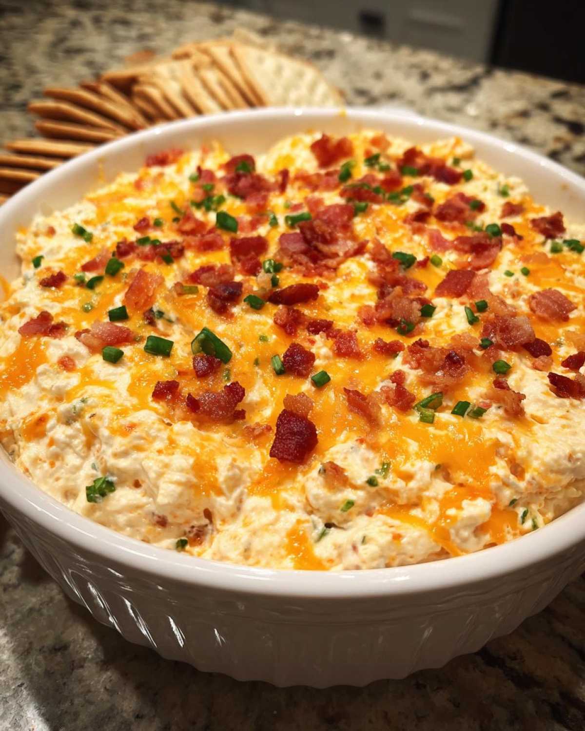 A close-up of creamy Million Dollar Dip topped with shredded cheddar cheese, crumbled bacon, and chopped chives, served with crackers.