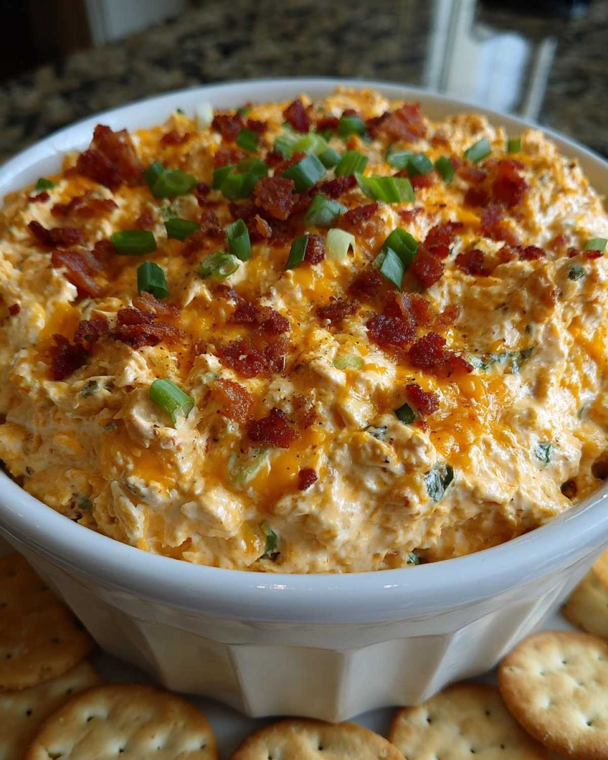Close-up of creamy Million Dollar Dip served in a white bowl, topped with crumbled bacon and green onions, surrounded by crackers.