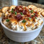 A close-up of creamy Million Dollar Dip topped with crumbled bacon, shredded cheddar, and chopped green onions, served with crackers.