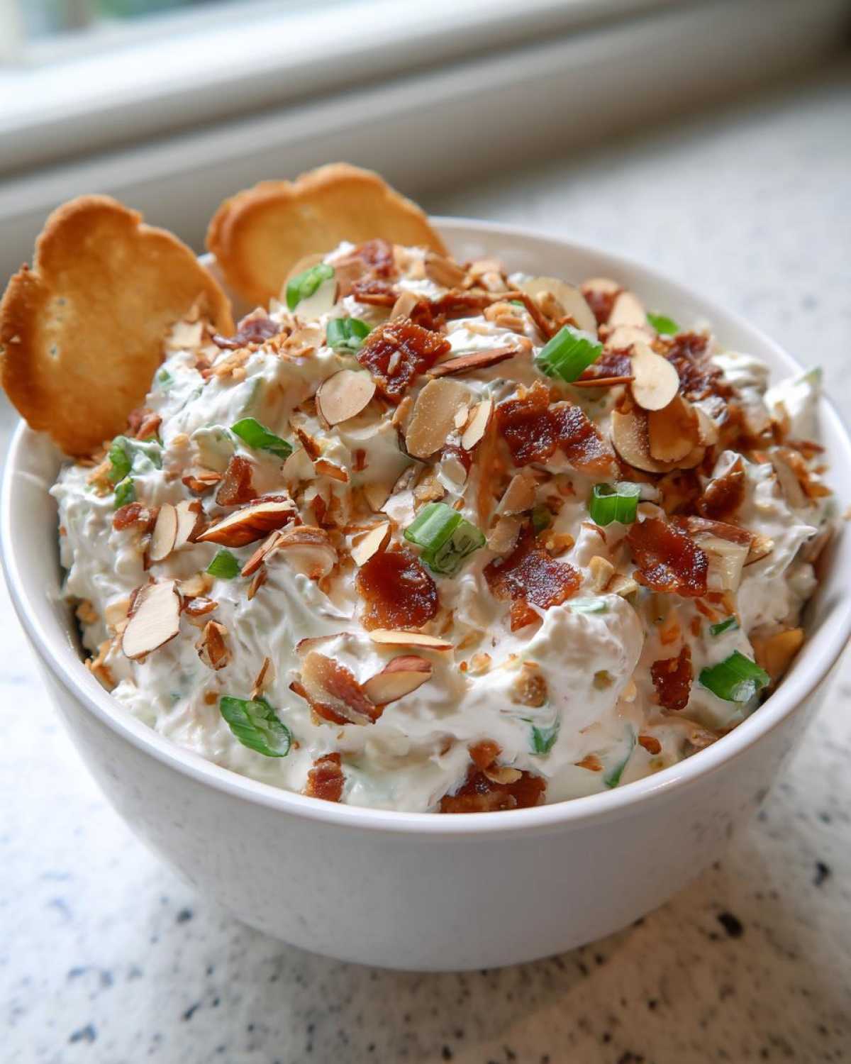 A white bowl filled with creamy Neiman Marcus dip, topped generously with bacon bits, sliced almonds, and green onions.