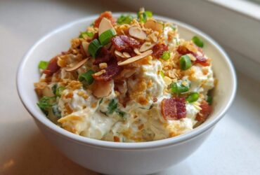 A white bowl filled with creamy Neiman Marcus dip, garnished generously with bacon bits, sliced almonds, and green onions.
