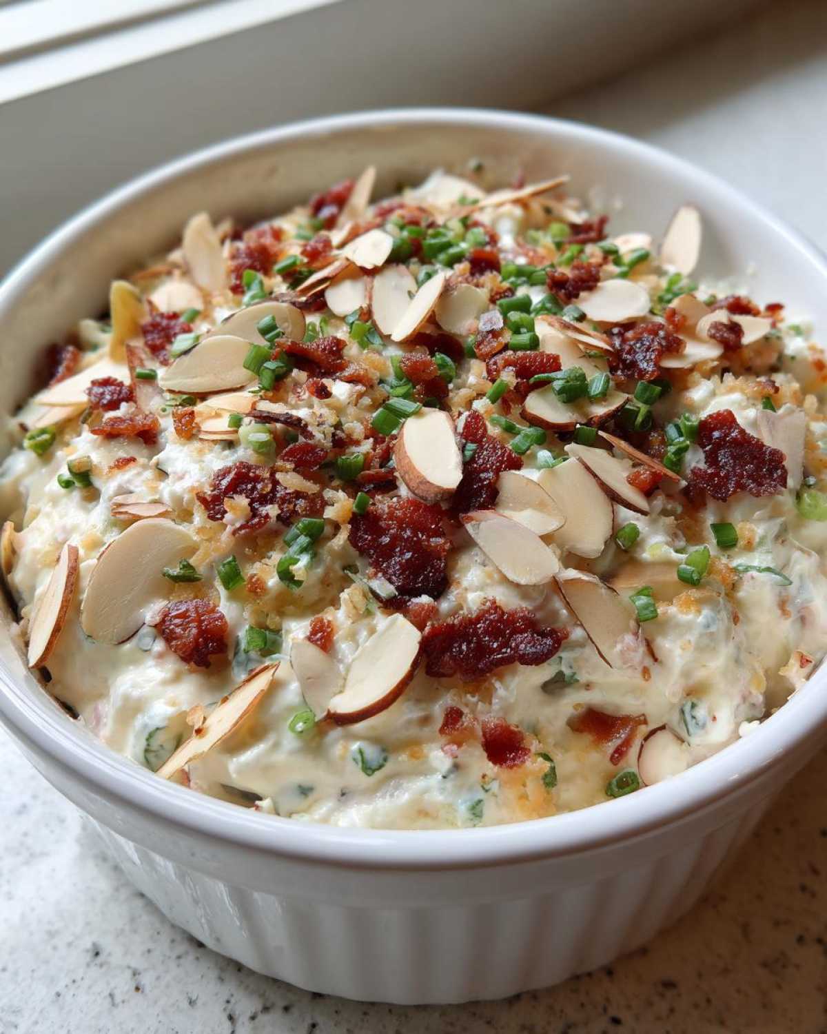 A close-up of creamy Neiman Marcus dip served in a white bowl, generously topped with sliced almonds, bacon bits, and chives.