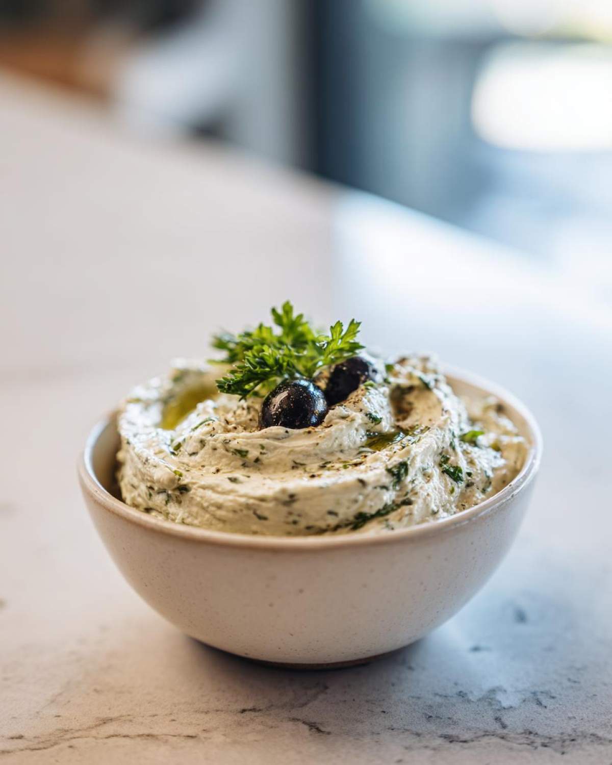 A close-up of a creamy, herb-flecked olive dip garnished with black olives and parsley.