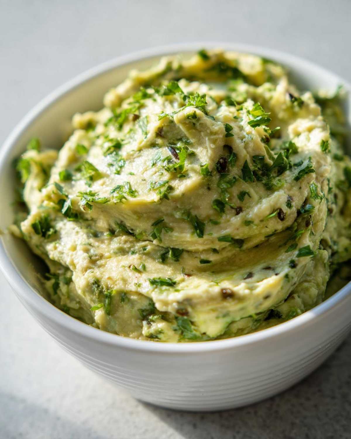 Close-up of creamy, light green olive dip topped generously with chopped fresh parsley in a white bowl.