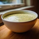 A creamy, pale green bowl of potato leek soup garnished with flecks of green herbs, sitting on a wooden board.