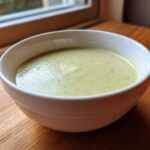 A close-up of a bowl of creamy, light green potato leek soup sitting on a wooden table near a window.