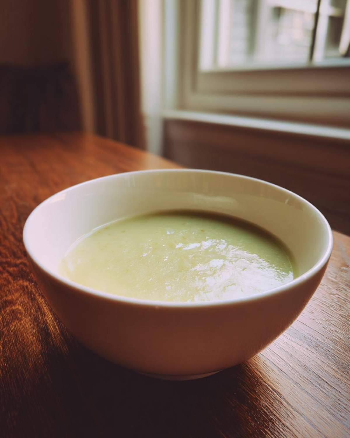A creamy bowl of smooth potato leek soup resting on a dark wooden table near a bright window.