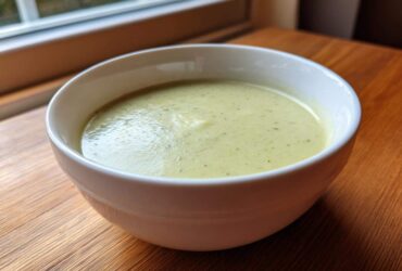 A close-up of a bowl of creamy, light green potato leek soup sitting on a wooden table near a window.