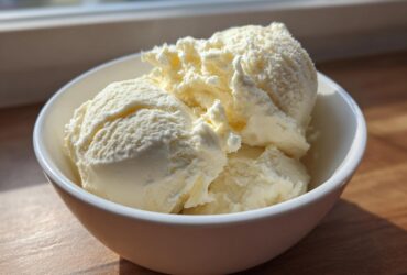 Three scoops of creamy vanilla-colored protein ice cream served in a small white bowl, highlighted by natural sunlight.