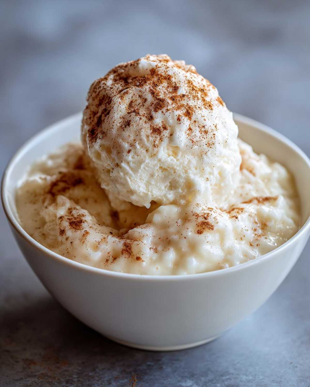 A white bowl filled with creamy rice pudding, topped with a scoop of cream and sprinkled generously with cinnamon.